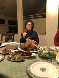 Janet's mom, an Asian woman wearing glasses, smiles and waves from behind a picture-perfect Thanksgiving turkey.