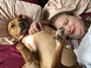 Katie - a white woman wearing red lipstick - and Ella - a tan dog with a white spot on her chest - are snuggling. Ella's belly is in the air, and Katie is smiling.