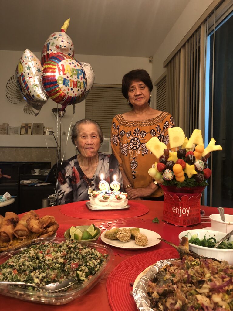 Myrla stands to the right of her mother on her 92nd birthday, with food and candles on a table in the foreground
