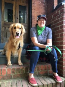 Erika, wearing a green bandana, sits on a brick stoop with a Golden Retriever who has a green leash on. They're both smiling at the camera.
