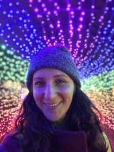 Tara, a white woman in a blue hat, is smiling at the camera. Behind her is a rainbow array of lights, and Tara's cheeks are glowing from the reflection.