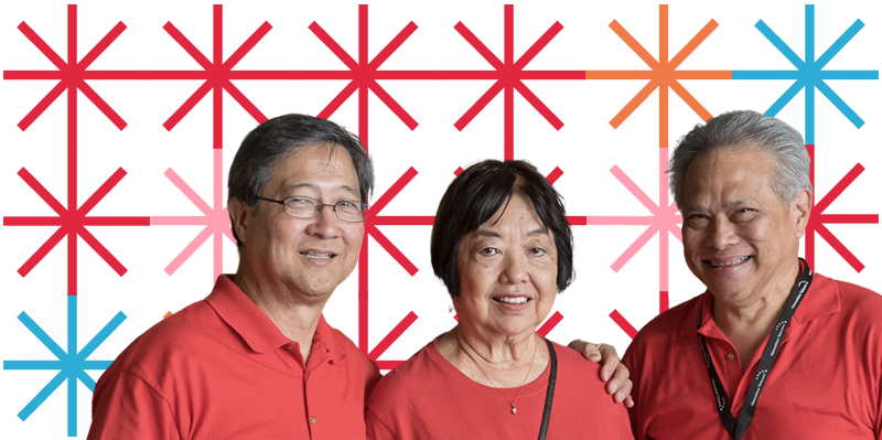 A group of three older adults smile while wearing matching red T-shirts and posing for the camera together.