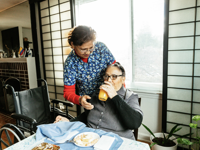 An older adult is enjoying a meal while an in-home care worker assists.