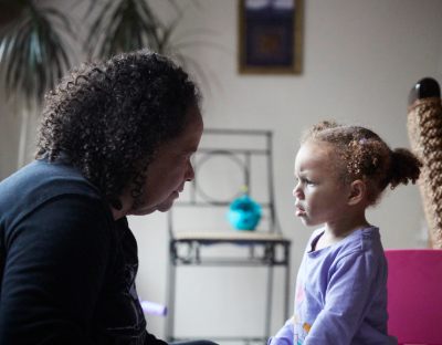 A toddler and an adult, both with dark curly hair, face each other and talk.