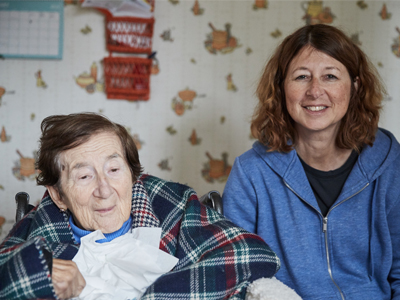 A family caregiver and an older adult, both with light skin and reddish brown hair, sit together and smile.