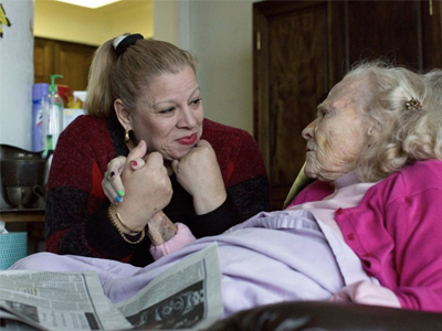 An in-home care worker holds hands and talks with an older person who is lying in bed.