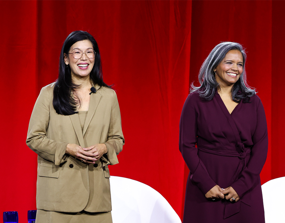 Ai-jen Poo and Sarita Gupta, founders of Caring Across Generations, address the crowd on stage at CareFest, smiling in front of a red velvet curtain.