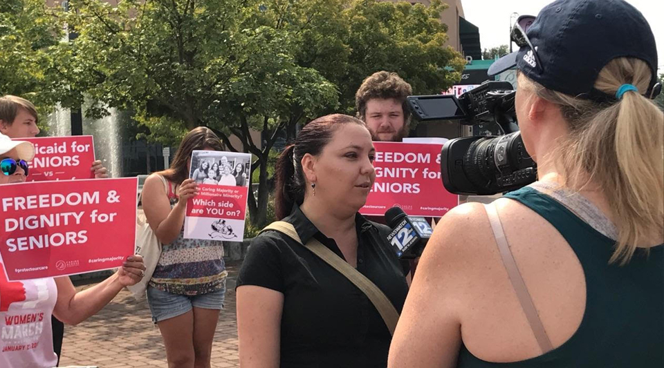 A television crew interviews an activist with light skin and a ponytail at a rally, with a group of people holding signs that read 