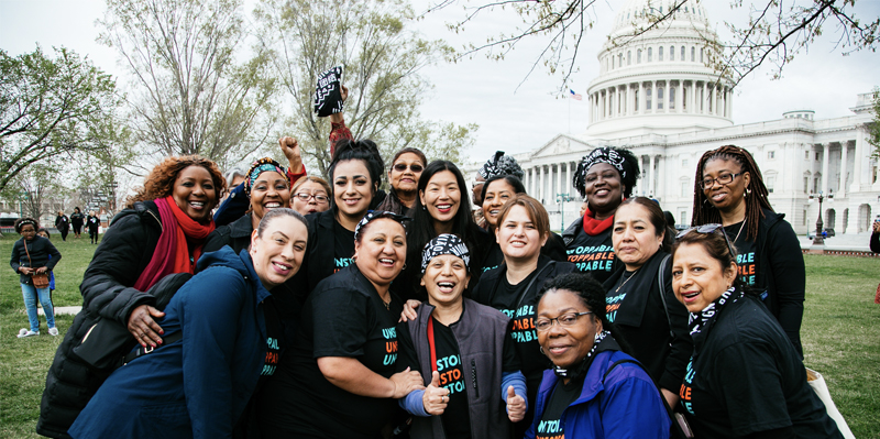 A group of people of varying ages and ethnicities and wearing matching T-shirts smile and celebrate outside the White House.