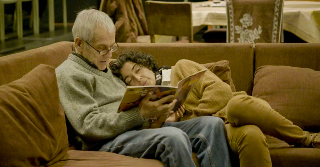 An image from the film 'The Eternal Memory' featuring a man and woman laying on a couch together reading a book.