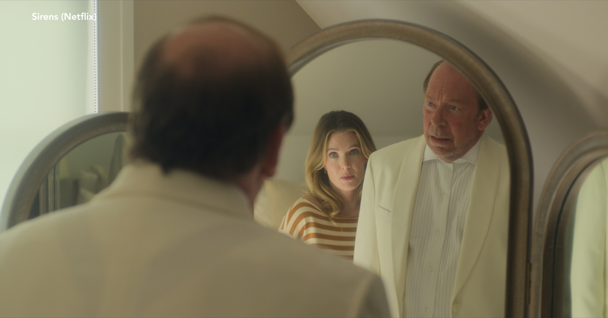 A man in a white suit looking into the mirror with his wife standing behind him.